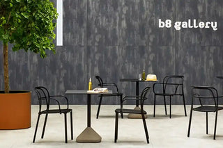 A modern dining or cafe seating arrangement featuring black metal chairs and square tables with concrete bases, set against a dark textured wall with the text 'b8 gallery' in white.